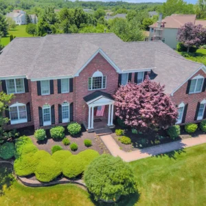 Aerial view of a two-story red brick colonial home with a new asphalt shingle roof, white columned front portico displaying an American flag, surrounded by lush green lawn, neatly trimmed shrubs, and a pink‑flowering tree.