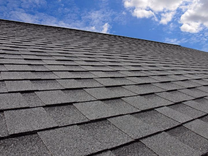 Close‑up of a sloped roof covered in dark gray asphalt shingles under a partly cloudy blue sky, labeled “Asphalt Shingle.”