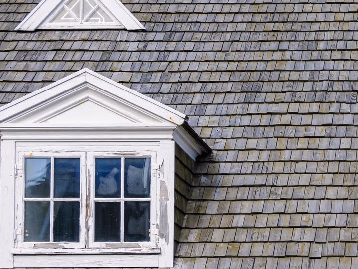 Close‑up of a roof covered in gray, weathered cedar shake shingles with a white dormer window.