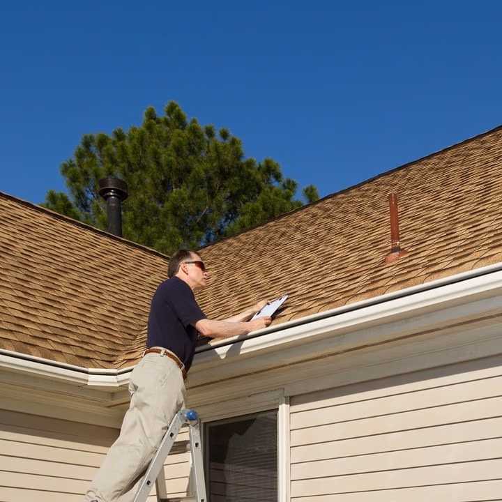 Riddick Roofing inspector on a ladder examining a brown asphalt shingle roof while holding a clipboard.