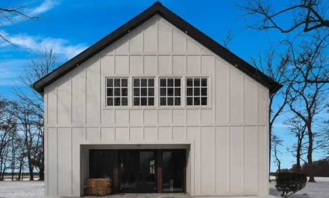 A white board-and-batten barn-style building with a steep black standing-seam metal roof, a row of multi-pane windows across the gable, large sliding glass doors at ground level, all set on a snowy yard framed by bare trees under a clear blue sky.